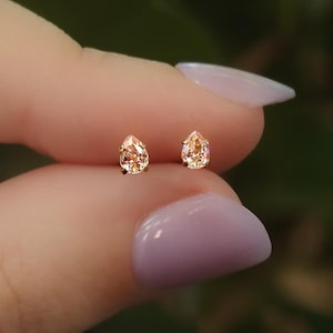May include: A pair of stud earrings featuring pear-shaped, light peach-colored gemstones set in gold-toned metal. The stones are faceted and reflect light, showcasing a clear, teardrop silhouette held by small prongs on each earring.