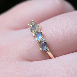 May include: A gold ring featuring three oval-cut labradorite stones with blue iridescence, accented by four small round clear stones set in prongs along the band. The stones are arranged in a row, showcasing the natural color play of the labradorite.