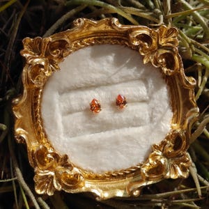 May include: A pair of pear-shaped orange gemstone stud earrings set in gold, displayed on a white velvet cushion inside a small, ornate, circular gold-colored frame with intricate scrollwork and decorative detailing along the edges.
