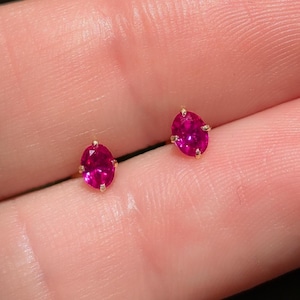 Peut inclure: Une paire de boucles d'oreilles à clous avec des pierres précieuses ovales d'un rouge rosé éclatant, montées sur des griffes en or. Chaque pierre facettée est maintenue par quatre petites griffes métalliques, offrant une couleur vive et saturée.