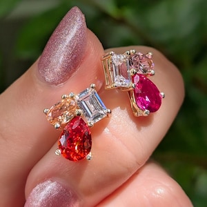 May include: Two gold stud earrings featuring a cluster of multi-colored gemstones. Each earring includes a clear emerald-cut stone, a pear-shaped red or orange stone, and a smaller oval-shaped stone in pink or peach, all set in gold metal prongs.