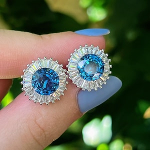 Puede incluir: Un par de pendientes de botón con una piedra preciosa azul brillante, grande y redonda en el centro, rodeada de piedras claras rectangulares más pequeñas. Los pendientes tienen un engaste de metal plateado y están sostenidos por una mano con esmalte de uñas azul.