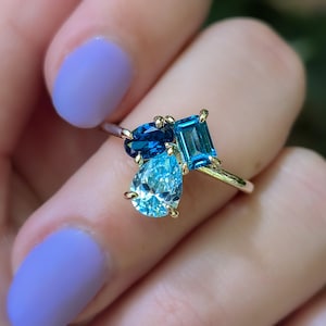May include: A gold ring featuring three blue gemstones: a pear-shaped light blue stone, a rectangular medium blue stone, and an oval dark blue stone. The ring is set against a blurred background.