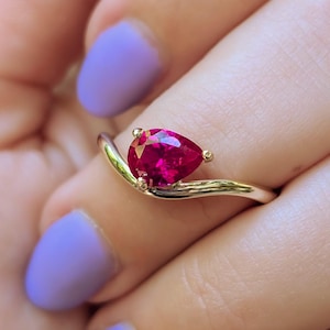 Pode incluir: Um anel de ouro com uma pedra preciosa de cor vermelho-rosada vibrante, lapidada em forma de pera e fixada por três garras. A banda tem um design liso, curvo e ondulado que segue o contorno da pedra em forma de lágrima, criando um perfil elegante.