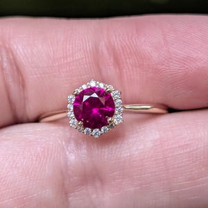 May include: A gold ring with a large, round, deep pink gemstone encircled by a halo of smaller, clear gemstones. The ring's band is a simple, thin gold. The gemstones are set in a hexagonal shape.