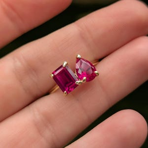 May include: A gold ring with two vivid, ruby-red gemstones. One stone is an emerald cut, the other a pear shape. The ring's band is slender and gold, with the gemstones held by gold prongs.