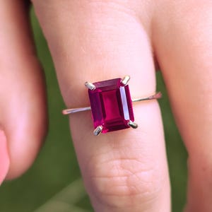 May include: A ring featuring a vibrant, rectangular, emerald-cut deep pink gemstone set in a simple, thin, polished silver-toned metal band. The stone is secured by four small prongs at each corner, showcasing a clear, faceted surface.