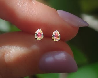 May include: A pair of small, teardrop-shaped stud earrings featuring iridescent, multi-colored stones that shift between pink, gold, and green hues. Each stone is secured in a simple metal prong setting, creating a delicate and reflective appearance.