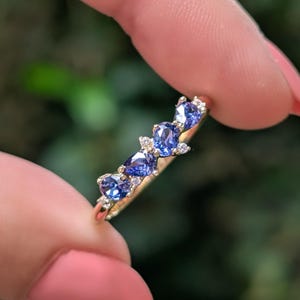 May include: A gold ring featuring five blue gemstones in a row, each with a different shape. Small white accent stones are set between the blue gems. The ring is held up against a blurred green background.