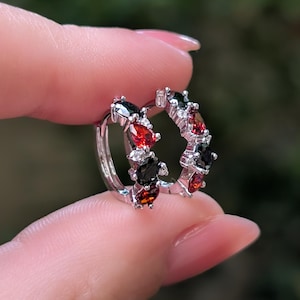 Può includere: Un paio di orecchini a cerchio color argento con pietre preziose alternate a forma di pera, di colore rosso e nero. I piccoli cerchi sono impreziositi da minuscole pietre chiare incastonate lungo la montatura in metallo.