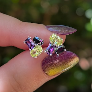 May include: A pair of stud earrings featuring a cluster of three faceted gemstones in silver-toned settings. Each earring contains one rectangular purple stone, one oval lime green stone, and one oval black stone arranged in a triangular formation.