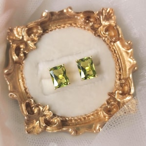 May include: A pair of rectangular, faceted lime green gemstone stud earrings set in gold prongs. The earrings are displayed on a white velvet ring holder insert within an ornate, circular, metallic gold frame with intricate scrollwork detailing.