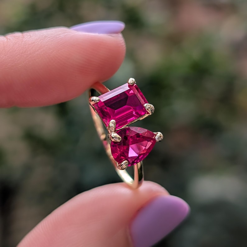 May include: A gold ring with two fuchsia gemstones, one rectangular and one pear-shaped. The ring is held up, showing the gemstones' colour and the ring's design. The background is blurred, highlighting the jewellery.