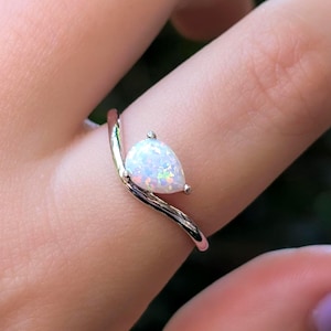 May include: A silver ring with a pear-shaped opal gemstone. The opal has a white base with iridescent flashes of color. The ring's band curves gracefully, and the gemstone is set with small prongs. The ring is shown on a finger.