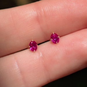 May include: A pair of small, oval-shaped, ruby-red gemstone stud earrings. The gemstones are set in a gold-colored metal. The earrings are displayed against a blurred background.