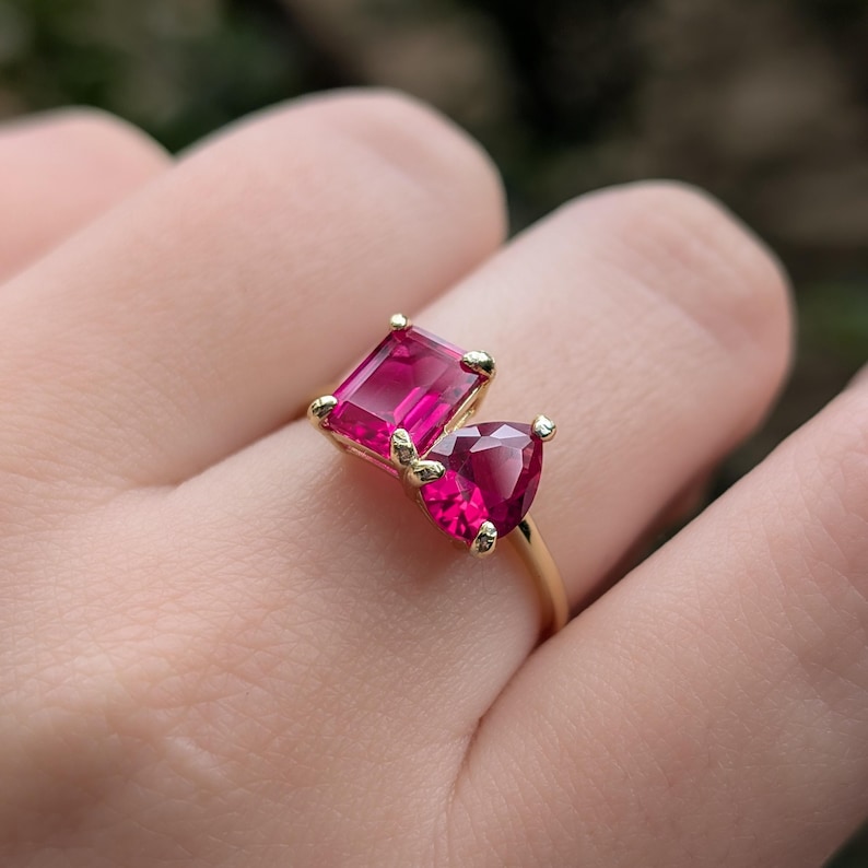 May include: A gold ring with two bright pink gemstones. One is rectangular, the other is heart-shaped. The ring has a plain, polished gold band, and the gemstones are held in place by small prongs.