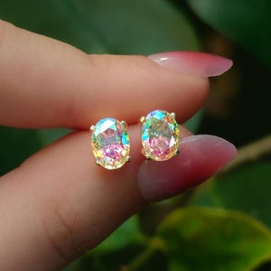 May include: A pair of oval-shaped stud earrings featuring iridescent, multi-colored faceted stones set in gold-toned metal prongs. The stones display a spectrum of shifting colors including pink, yellow, and blue hues with a high-gloss finish.