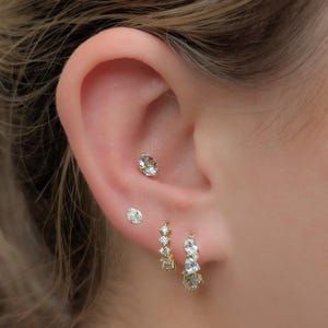 May include: Close-up of an ear with multiple earrings. Three small, clear, oval-shaped stud earrings and a pair of gold hoop earrings with clear, oval-shaped stones. The earrings are set against the skin.