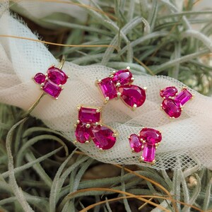 Peut inclure: Ensemble de bijoux en or orn&eacute;s de pierres pr&eacute;cieuses roses vives. L'ensemble comprend une bague et plusieurs paires de boucles d'oreilles, chacune pr&eacute;sentant un groupe de pierres de formes diff&eacute;rentes. Les bijoux sont pr&eacute;sent&eacute;s sur un tissu blanc sur fond de feuillage vert.