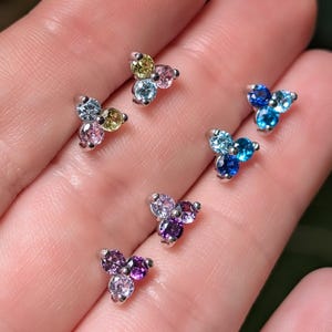 May include: A collection of small, silver-toned stud earrings. Each earring features three round, colorful gemstones in various shades of blue, purple, pink, and yellow. The earrings are displayed on a light skin tone.