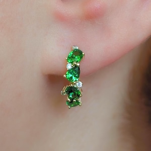 May include: A gold-toned earring with a series of vibrant green gemstones and small, clear accent stones. The gemstones are oval and pear-shaped, arranged in a curved line. The earring is set against a blurred background.