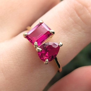 May include: A gold ring featuring two vibrant red gemstones, one in a rectangular emerald cut and the other in a teardrop pear cut. The stones are set in a polished gold band with delicate prongs securing each gem in this dual-stone design.