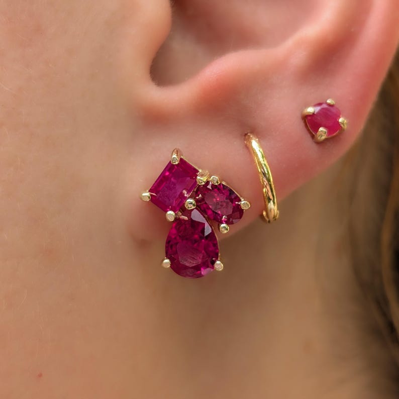 Peut inclure: Gros plan sur une oreille orn&eacute;e de trois boucles d'oreilles en or. Une boucle d'oreille pr&eacute;sente un groupe de trois pierres pr&eacute;cieuses de couleur rubis de formes carr&eacute;es, ovales et en goutte. Un petit clou d'oreille en rubis et une cr&eacute;ole en or sont &eacute;galement visibles.