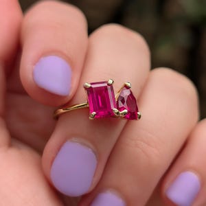 May include: A gold ring featuring two vibrant, rectangular and pear-shaped, ruby-red gemstones. The ring's band is a delicate gold, and the gemstones are set in a gold bezel. The ring is held by a person with purple nail polish.