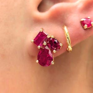 Peut inclure: Gros plan sur une boucle d'oreille en grappe avec une monture dor&eacute;e et trois pierres pr&eacute;cieuses de couleur rubis. Une petite cr&eacute;ole dor&eacute;e et une boucle d'oreille &agrave; clou en rubis sont &eacute;galement visibles. Les boucles d'oreilles sont pos&eacute;es sur un fond de teint.