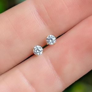 May include: A pair of small, round stud earrings. Each earring features a clear, sparkling gemstone set in a gold-colored metal. The earrings are displayed against a soft, blurred background.