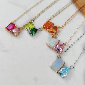 May include: A collection of gemstone necklaces with gold and silver chains. Each necklace features two colorful gemstones in various shapes, including rectangular, pear, and heart-shaped stones. The gemstones are in shades of pink, green, orange, and blue.