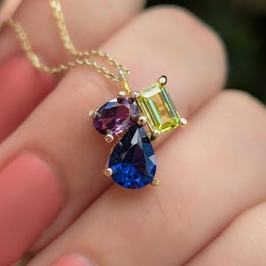 May include: A gold necklace featuring a pendant with three colorful gemstones. The pendant has a pear-shaped blue gemstone, a heart-shaped purple gemstone, and a square-cut green gemstone. The necklace is a delicate piece of jewelry.