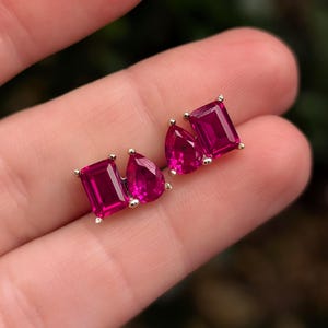 Op de afbeelding: Stud oorbellen met een rechthoekige en een peervormige, levendige magenta edelsteen gezet in goud. De oorbellen worden gepresenteerd tegen een neutrale achtergrond, waarbij de kleur en het ontwerp van de sieraden worden benadrukt.