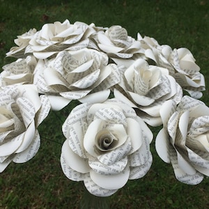 May include: A bouquet of handcrafted paper roses made from recycled book pages with visible printed text. Each flower features multiple layered petals curled into a bloom shape, showcasing black typography on off-white paper throughout the arrangement.