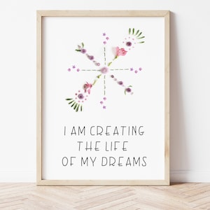 May include: A white framed print with a floral design in shades of pink and purple. The design is a stylized star with the text "I AM CREATING THE LIFE OF MY DREAMS" below it.
