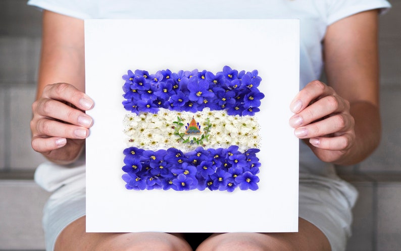 May include: A white card with a blue and white flower arrangement depicting the flag of El Salvador. The card is being held by a person's hands.