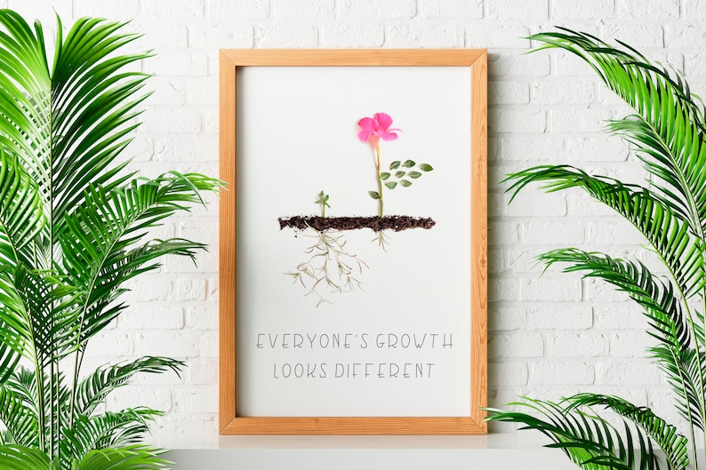 Op de afbeelding: Een ingelijst print met een witte achtergrond waarop een roze bloem en twee groene bladeren te zien zijn die uit de grond groeien. De wortels van de planten zijn zichtbaar onder de grond. De tekst "Everyone's growth looks different" is onder de afbeelding gedrukt.