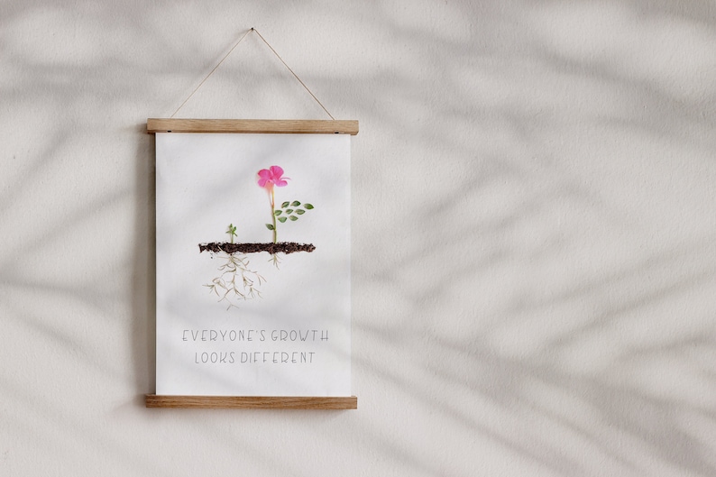 Op de afbeelding: Een ingelijst print met een roze bloem, groene bladeren en bruine wortels die uit de grond groeien. De tekst "Everyone's growth looks different" is onder de afbeelding gedrukt.
