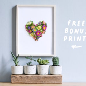 Op de afbeelding: Een ingelijste print van een hartvorm gemaakt van geperste bloemen in tinten roze, paars en geel. De print wordt weergegeven op een muur met een plank eronder met vijf potplanten.