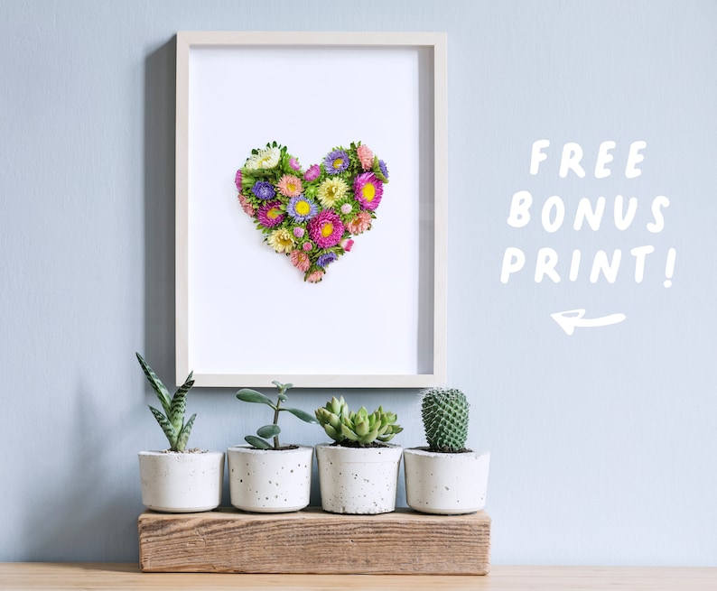 Op de afbeelding: Een ingelijste print van een hartvorm gemaakt van geperste bloemen in tinten roze, paars en geel. De print wordt weergegeven op een muur met een plank eronder met vijf potplanten.