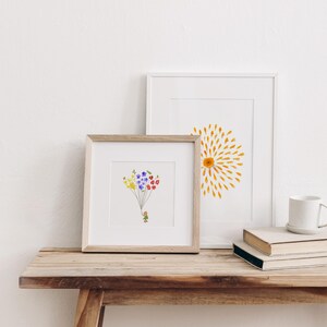 May include: Two framed prints on a wooden table. The print on the left features a small figure holding a bunch of colorful flower balloons. The print on the right features a yellow sunburst design.