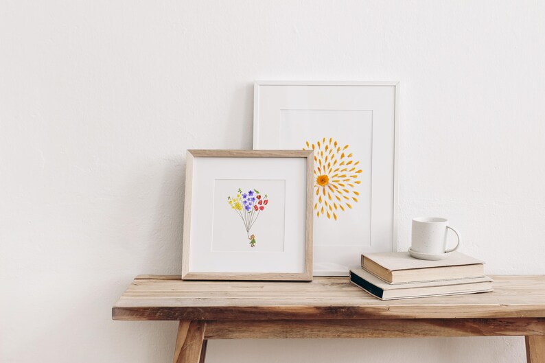 May include: Two framed prints on a wooden table. The print on the left features a small figure holding a bunch of colorful flower balloons. The print on the right features a yellow sunburst design.