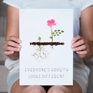 Op de afbeelding: Een witte kaart met een roze bloem en groene bladeren die uit bruine aarde groeien. De tekst "Everyone's growth looks different" staat op de kaart gedrukt.