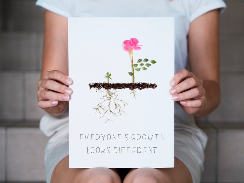Op de afbeelding: Een witte kaart met een roze bloem en groene bladeren die uit bruine aarde groeien. De tekst "Everyone's growth looks different" staat op de kaart gedrukt.