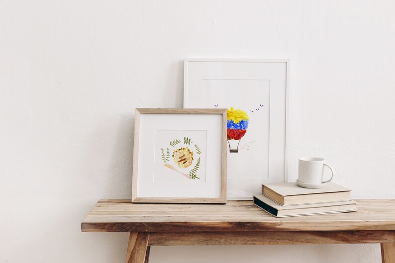 May include: Two framed floral art prints on a wooden table. The print on the left features a hedgehog made of flowers and leaves. The print on the right features a hot air balloon made of flowers.