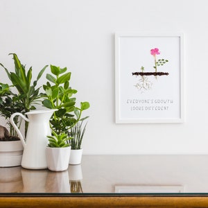 Op de afbeelding: Een witte ingelijste print met een roze bloem en groene bladeren die uit de grond groeien. De tekst "Everyone's growth looks different" is onder de afbeelding gedrukt.
