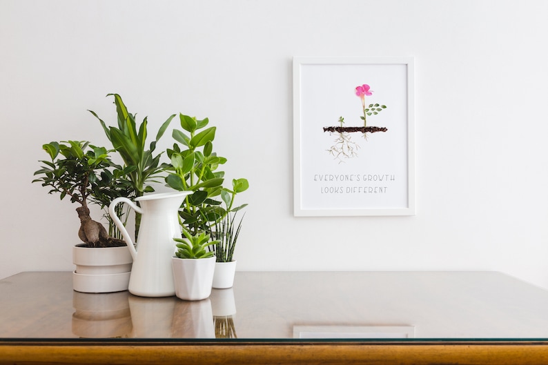 Op de afbeelding: Een witte ingelijste print met een roze bloem en groene bladeren die uit de grond groeien. De tekst "Everyone's growth looks different" is onder de afbeelding gedrukt.