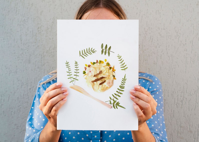 May include: A white card with a pressed flower arrangement in the center. The arrangement is a circle of yellow and white flowers with a brown stem and green leaves. A wooden spoon is placed below the flower arrangement. The card is surrounded by a border of green leaves.