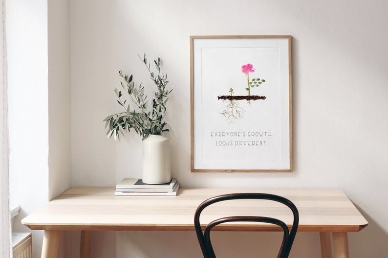 Op de afbeelding: Een ingelijst print met een roze bloem en groene bladeren die uit de grond groeien. De tekst "Everyone's growth looks different" is onder de afbeelding gedrukt.