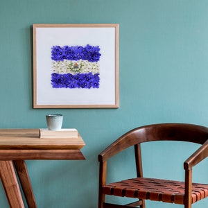 May include: A framed print of a flag made of purple and white flowers. The flag is displayed on a light blue wall above a wooden table and chair.